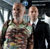 Two pixelated figures stand side-by-side; one wears a camouflage-patterned shirt, the other a dark suit with a white collar, against a blurred background with light and dark tones.