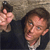 A pixelated figure in dark clothing holds a small object near their face against a textured background.