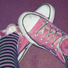 Close-up of pink canvas sneakers with white laces and black trim, worn with black-and-white striped leggings against a purple background.