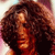 A pixelated close-up of a person with voluminous curly brown hair, wearing a dark top against a light background with pink and blue tones.