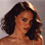 A pixelated close-up of a person with dark curly hair, light brown skin, and a neutral expression against a gray background.
