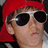 Pixelated close-up of a person wearing dark sunglasses and a red head covering, with visible ear and partial face.
