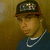 Pixelated static image of a person wearing dark cap, blue shirt, and silver chain necklace against brown background.