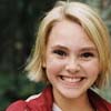 A static image of a person with short blonde hair wearing a red jacket with white stripes over a white shirt, set against a blurred green background.