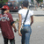 Two people walking side-by-side on a street, one in a maroon shirt and red cap, the other in a white shirt and blue jeans with a black strap across the back.