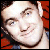 Pixelated close-up of a person's face with light skin, dark eyebrows, and a slight smile, set against a red and dark background.