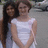 Two women in white dresses stand side-by-side outdoors, one with long dark hair and the other with short brown hair, both smiling.