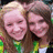 Two people with long brown hair and light skin pose closely together, both wearing green-accented tops against a blurred background.