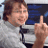 Pixelated image of a person with brown hair wearing a white shirt, holding up a single finger in front of a blue and dark background.