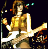 A person with short brown hair plays a white electric guitar on stage, wearing a gold sleeveless top and dark shorts under dim lighting.