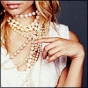 A close-up of a person wearing multiple gold chain necklaces over a white sleeveless top, with a hand positioned near the chest.