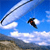 Pixelated static image of a parachutist in mid-air against a blue sky with clouds and distant terrain.