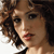 Pixelated close-up of a person with curly brown hair, light skin, and red lips, wearing a black and white garment against a light background.