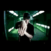 A person in a black shirt and dark cap performs hand gestures against alternating green tunnel and crowd backgrounds with bright lights.