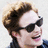 Pixelated close-up of a person with dark brown hair styled in a bun, wearing black sunglasses and a dark blue top, smiling with visible teeth.