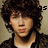 Pixelated static image of a person with dark curly hair, light skin, and a black jacket over a white shirt against a beige background.