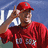 A person in a red baseball uniform and cap adjusts their cap with both hands, standing before a blue background with partial white text.