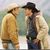 Two figures in period clothing face each other against a blurred background, one wearing a light-colored jacket and hat, the other in a dark jacket and wide-brimmed hat.