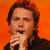 Pixelated image of a person with brown hair holding a microphone against an orange background.