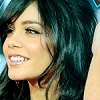 A close-up of a person with long dark hair smiling, showing teeth, against a blurred background.