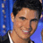 A person with short dark hair and a black jacket over white shirt smiles against blue background with light spots.