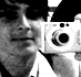A person holds a silver digital camera with a circular lens and black buttons, wearing a white collared shirt against a blurred background.