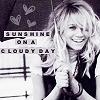 A grayscale image of a smiling woman with long blonde hair holding a microphone, overlaid with text reading "SUNSHINE ON A CLOUDY DAY" and heart symbols in the background.