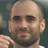 Pixelated static image of a bald man with stubble and dark eyebrows, wearing a light-colored shirt against a blurred background.