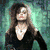 A woman with long brown hair and a black top stands against a greenish background with a white necklace.