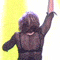 A woman with shoulder-length brown hair sings into a microphone while raising one arm, set against a colorful striped background.