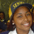 A pixelated close-up of a person wearing a dark cap with yellow tassels, a white shirt, and earrings, with a blurred background.
