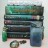 Pixelated static image of stacked books with green and blue spines, a small circular object below, and a light background.