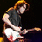 Pixelated image of a person with light brown hair playing a white electric guitar against dark background.