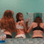 Three women in swimwear stand side-by-side against a light blue background, with the central figure's arm raised.