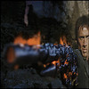 A pixelated image shows a person holding a large, dark firearm with visible orange muzzle flash against a dark, rocky background.