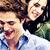 Two people in close proximity, one with dark hair and a white shirt, the other with light hair and a dark top, both smiling against a blurred green background.