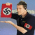 A person in a dark polo shirt holds a red square with a black and white swastika symbol, extending their arm forward while standing against a light blue background.