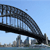 Pixelated image of a steel arch bridge spanning water with city buildings in the background under a clear blue sky.