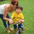 A child in a yellow shirt and blue shorts rides a bicycle with an adult behind, both on grass.