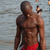 A shirtless man in red shorts stands in shallow water, his head turning from left to right