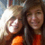 Two people in close proximity, one wearing orange clothing, the other in dark clothing with a light-colored collar, against a blurred background with bright light on the left.