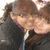 Two people in dark jackets with light zippers pose closely together, one with brown hair tied back, against a light background.