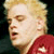 Pixelated close-up of a person with light hair wearing a red garment with a white star, against a dark background.