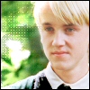 A close-up of a person with light blonde hair wearing a white collared shirt and dark tie against a blurred green background.
