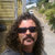 Pixelated static image of a person with long dark hair wearing dark sunglasses, standing outdoors with blurred green and brown background elements.