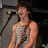 A person in a sleeveless shirt sings into a microphone with a dark background and stage lighting.