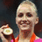 A person with short blonde hair holds a round object in their right hand, wearing an orange necklace and pink top against dark background.