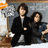 Two figures in dark clothing sit side-by-side against a muted background with an orange and white object on the left.