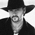 Black-and-white pixelated icon of a person wearing a wide-brimmed hat and dark coat with visible facial features including eyes, nose, mouth, and beard.