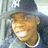 Pixelated image of a smiling character with light brown hair, wearing a dark blue top and white collar, set against a blurred background.